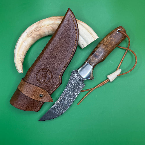 A fixed-blade knife with a patterned steel blade, a brown wooden handle, and a cord lanyard, displayed next to its brown leather sheath on a solid green background. A curved ivory horn is positioned behind the sheath.